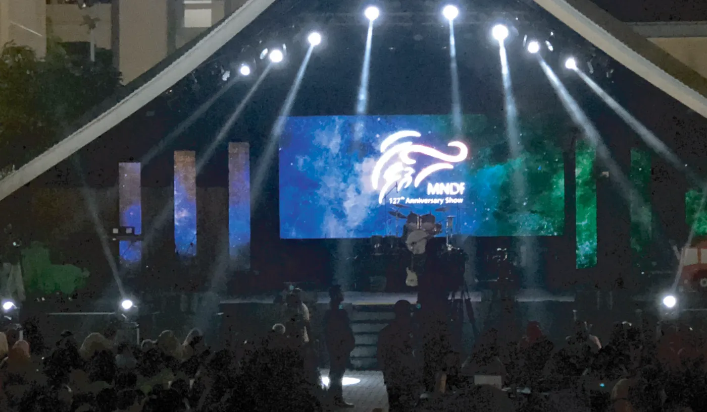 Stage with moving-head lights and an LED screen showing the MNDF 127th Anniversary Show logo, audience in the foreground